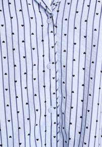 Lyseblå skjorte med knapper, prydet med lodrette lilla striber og små sorte hjerte-mønstre. Fremstillet af blødt stof med en glat tekstur.