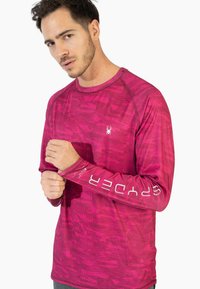 Pitkähihaisi urheilupaita elinvoimaisessa pinkissä, jossa on teksturoitu kuvio. Rinnassa logo ja hihoissa painettu brändinimi.