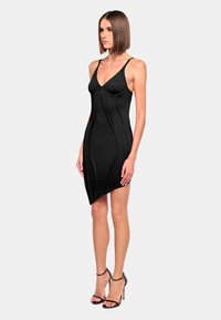 Abito nero aderente con spalline sottili, cuciture contour e orlo asimmetrico. Realizzato in tessuto liscio con una leggera lucentezza. Abbinato a tacchi neri.