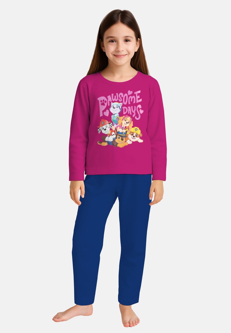 Ragazzina che indossa una maglietta lunga rosa con cani dei cartoni animati e la scritta "Pawsome Days", abbinata a pantaloni blu, in piedi a piedi nudi.