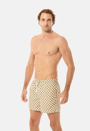 Paitaansa riisunut mies, jolla on beige- ja valkokuviolliset uimalasit, joissa on nyörikiristys, seisomassa yksiväristä vaaleaa taustaa vasten.