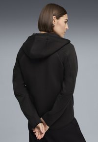 Donna con capelli corti castani che indossa una felpa nera con cappuccio, vista di profilo con le mani intrecciate dietro la schiena su uno sfondo grigio.