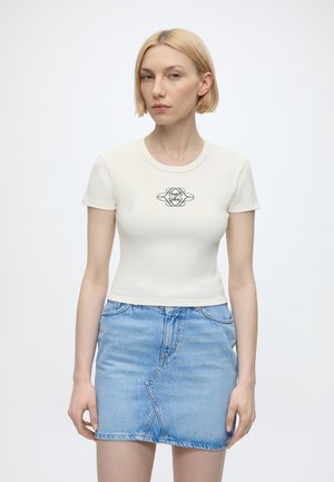 Femme aux cheveux blonds courts portant un T-shirt blanc ajusté avec un logo et une mini-jupe en jean bleu clair, debout devant un fond blanc.