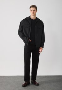 Veste noire à boutons avec deux poches avant, assortie à un t-shirt noir et un pantalon noir. Chaussures bordeaux brillantes. Design simple, coupe moderne.