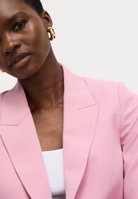 Blazer ajusté rose clair à revers, porté sur un haut blanc, agrémenté de grandes boucles d'oreilles en or. Texture de tissu lisse, vue en gros plan.