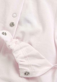 Vêtement en coton rose clair avec des fermetures à pression argentées, mettant en avant un détail de manche froncé et une couture visible le long de l'ourlet.