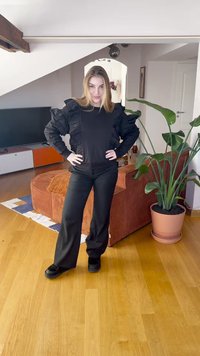 Svart outfit med en texturerad, volangprydd topp och löst sittande byxor. Svarta skor. Inomhusmiljö med en växt och möbler i bakgrunden.
