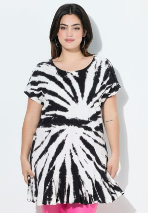 Kvinde med mørkt hår iført sort og hvid tie-dye kjole med korte ærmer og flæsekant, stående foran en hvid baggrund.