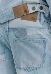 Jean en denim patchwork bleu clair avec grande poche arrière et étiquettes de marque en cuir marron sur la ceinture, porté avec une veste en denim assortie.