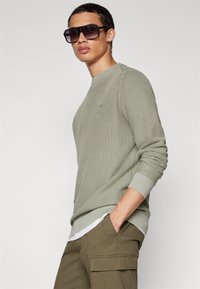 Grüner, strukturierten Pullover mit Rundhalsausschnitt, langen Ärmeln und gerippten Bündchen, kombiniert mit olivfarbenen Cargo-Hosen und übergroßen, dunklen Sonnenbrillen.