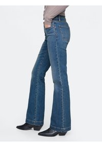 Kvinna i blå bootcut-jeans, svarta spetsiga klackstövlar och en ljusbrun långärmad topp, med en hand i fickan.