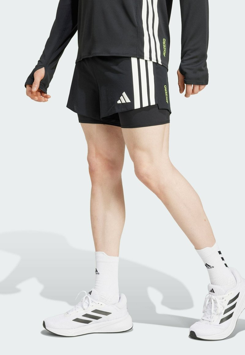 Mustat urheilushortsit, joissa on valkoiset pystysuorat raidat, yhdistettynä valkoisiin sukkiin ja kenkiin. Huomionarvoisia yksityiskohtia ovat Adidas-logo ja brändäys.