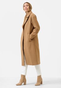 Lichtbruin wollen jas met een insnoering, lange mouwen en zijzakken, gecombineerd met beige lakleren enkellaarsjes en witte broeken.