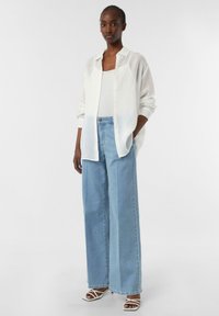 Weißes Leinen-Hemd mit Knöpfen über einem hellen Tanktop, kombiniert mit weit geschnittenen, hellblauen Jeans und weißen Sandalen mit Riemchen. Schlicht, entspannter Schnitt.