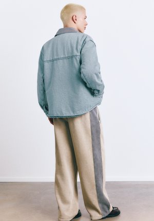 Osoba o krótkich blond włosach w wyblakłej, niebieskiej, oversize'owej kurtce i beżowych szerokich spodniach z szarymi paskami po bokach, stojąca w pomieszczeniu.