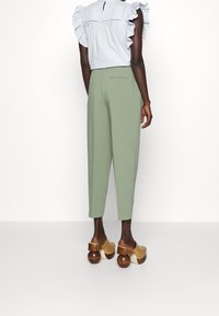 Groene tapse broek met een gladde textuur, zijzakken, gecombineerd met een lichtblauw blouse met gerimpelde mouwen en klompen met houten hakken.