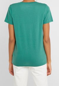 Persona che indossa una semplice maglietta teal a maniche corte e pantaloni bianchi, vista da dietro su uno sfondo neutro.