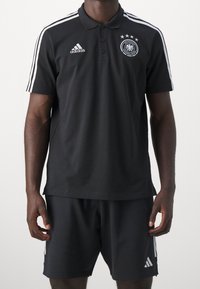 Černé polo tričko s bílými pruhy Adidas na ramenou, logo na hrudi, zdobeno německým národním emblémem a čtyřmi hvězdami nad ním. V kombinaci s černými šortkami.
