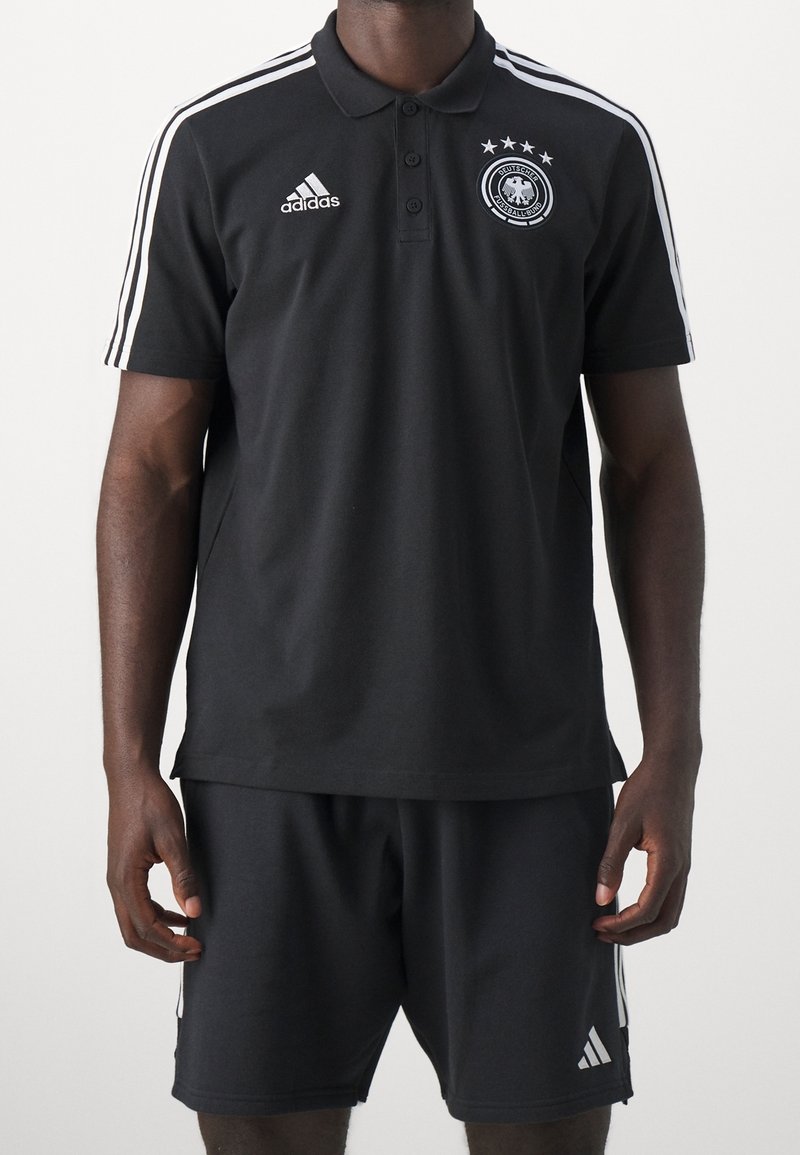 Černé polo tričko s bílými pruhy Adidas na ramenou, logo na hrudi, zdobeno německým národním emblémem a čtyřmi hvězdami nad ním. V kombinaci s černými šortkami.