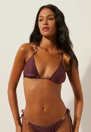 Frau mit langen dunklen Haaren, die einen metallisch-lila String-Bikini mit goldenen Verschlussdetails trägt und vor einem schlichten hellen Hintergrund steht.