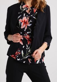 Veste noire à manches trois-quarts superposée à un chemisier floral avec des lys rouges et des feuilles vertes sur fond noir.