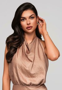 Haut en tissu drapé de couleur brun clair, avec un design fluide et texturé. Le mannequin a de longs cheveux foncés et porte une boucle d'oreille en or.