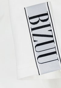 Biała tkanina z wszytą szaro etykietą z czarnym tekstem. Etykieta ma pionową orientację i teksturowaną powierzchnię.