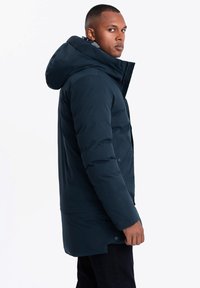 Marinblå puffjacka med en stor huva, frontknäppning och textilstruktur. Har en rak passform och design i höftlängd.