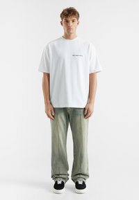 Hvit bomulls T-skjorte med korte ermer og rund hals, med svart tekst "don't waste culture" på forsiden. Kombinert med falmede grønne jeans og svarte sneakers.