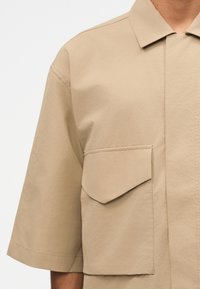 Chemise beige à manches courtes avec une grande poche à rabat sur le devant et un col pointu, portée par une personne visible de la poitrine aux épaules.