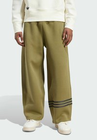 Olivově zelené tepláky vyrobené z měkké tkaniny, s �širokými nohavicemi, postranními švy a třemi černými pruhy na spodní části nohavic.