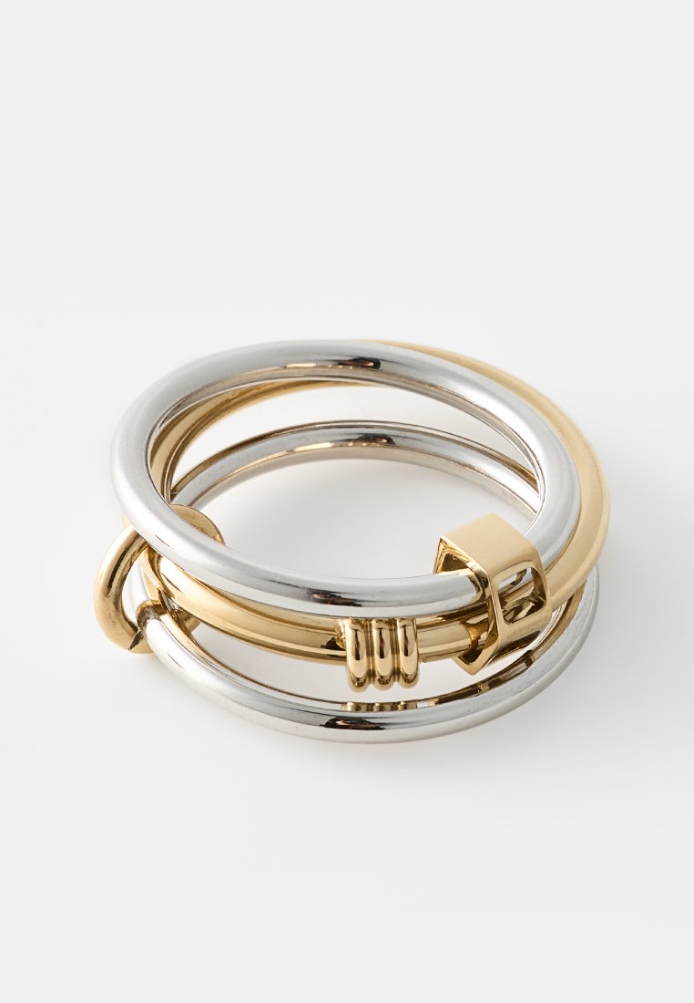 Drei verschlungene Ringe in Silber- und Goldtönen, mit einer glatten, polierten Oberfläche und einem markanten Schnappverschluss-Detail an einem Ring.