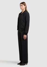 Femme debout de profil portant une veste bomber matelassée noire, un pantalon noir à jambes larges et des bottines noires, sur un fond uni.