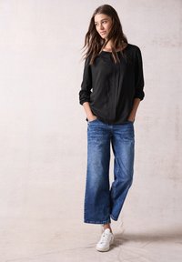 Maglietta nera a maniche lunghe con patta frontale, abbinata a jeans blu a gamba larga e sneakers bianche. Design semplice e vestibilità ampia.