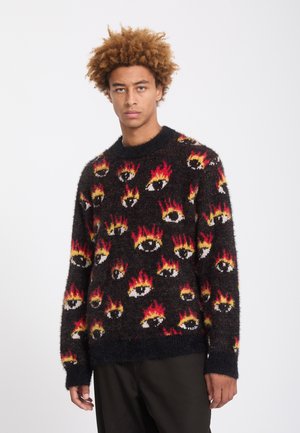 Jeune homme aux cheveux bouclés portant un pull noir duveteux recouvert de motifs colorés d’yeux et de flammes, debout devant un fond clair uni.