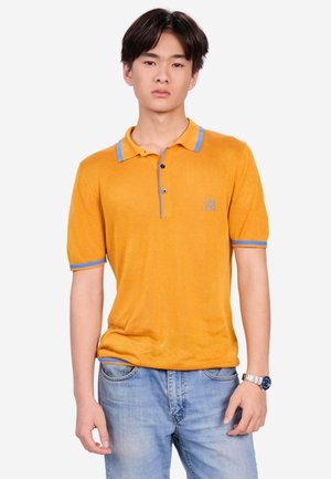 Junger Mann trägt ein senfgelbes Poloshirt mit blauem Rand und hellblaue Jeans, steht mit neutralem Gesichtsausdruck vor einem einfarbigen Hintergrund.