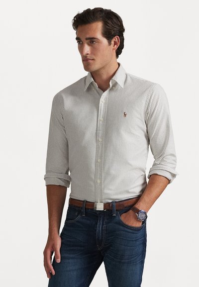 Camisa de rayas blanca y gris claro, con un cuello clásico y las mangas remangadas. Combinada con jeans azul oscuro y un cinturón marrón.
