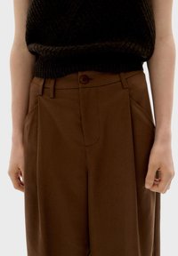 Bruine op maat gemaakte broeken van soepel fabric, met een knoopsluiting, zijzakken en een rechte snit. Detail op de tailleband zichtbaar.