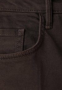 Gros plan d'une poche de jean en denim brun foncé avec coutures visibles et passant de ceinture sur tissu texturé.