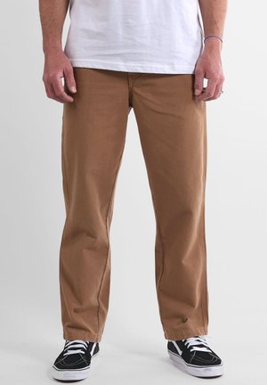 Pantalon chino en coton marron avec une coupe droite, doté de poches avant et de coutures renforcées. Porté avec des chaussures noires et blanches.