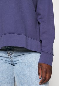 Personne portant un sweat-shirt violet à manches longues et un jean denim bleu clair, avec la main droite détendue le long du corps.
