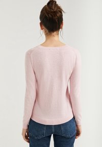 Woman with brown hair in a bun wearing a light pink long-sleeve sweater and blue jeans, shown from the back against a plain background.