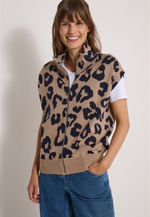 Kvinde iført en ærmeløs vest med lynlås med tan- og sort leopardprint, hvid kortærmet skjorte og blå jeans, smilende med den ene hånd i lommen.