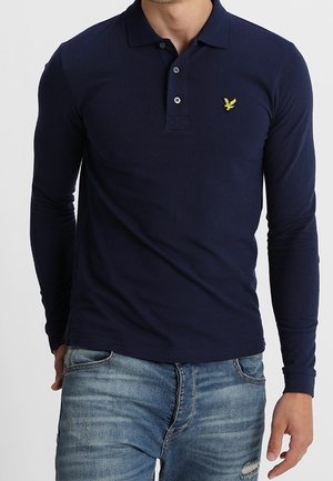 Marineblå langærmet polo-shirt med en krave med tre knapper og et gult ørnemblem på venstre bryst; struktureret stof. Brugt med jeans.