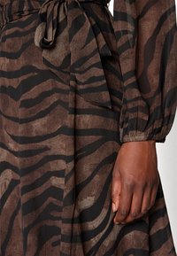 Robe rayée zèbre marron et noire avec une ceinture à la taille et de longues manches bouffantes, portée par une personne à la peau foncée.