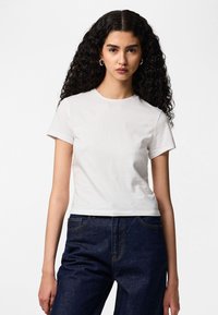Giovane donna con lunghi capelli ricci che indossa una t-shirt bianca corta semplice e jeans a vita alta di colore blu scuro, di fronte.