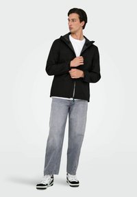 Giacca nera con cerniera e cappuccio grigio, caratterizzata da una texture liscia. Abbinata a jeans grigio chiaro e sneaker bianche e nere.