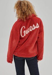 Rode jas van nep bont met wit "Guess"-logo op de achterkant, met een losse pasvorm en lange mouwen, gecombineerd met donker denim.