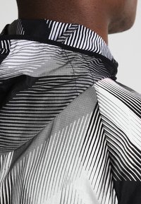 Lett jakke med et dynamisk sort og hvidt stribet mønster, lavet af en blanding af glatte og teksturerede stoffer. Hætte-design.