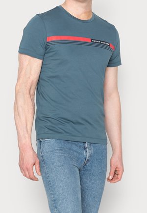 Mand iført en turkis Tommy Hilfiger t-shirt med røde og sorte striber og lyseblå jeans, stående mod en ensfarvet hvid baggrund.